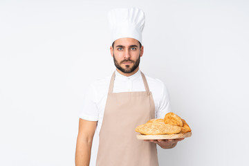 Male baker holding a table with several breads isolated on white background keeping arms crossed