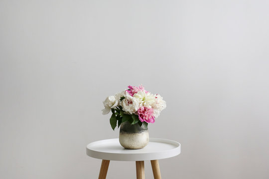 Close up shot of beautiful decorative glass vase with bouquet of peonies on white coffee table on foreground and blank wall with a lot of copy space for text on background.
