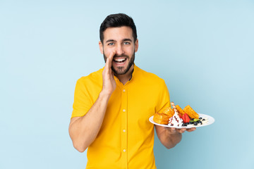 Caucasian man holding waffles isolated on blue background shouting with mouth wide open