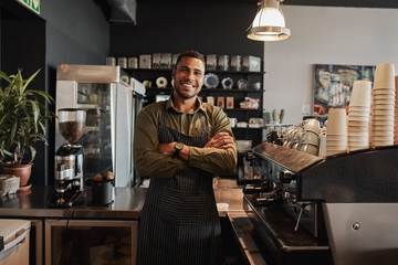 Portrait of a smiling confident young waiter standing at the cafe counter - obrazy, fototapety, plakaty
