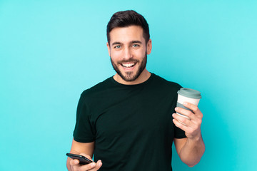 Caucasian handsome man isolated on blue background holding coffee to take away and a mobile