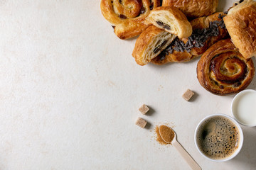 Variety of traditional french puff pastry buns with rasin and chocolate, croissant with paper cup of coffee and milk, recycled wooden spoon of cane sugar over white texture background. Flat lay, space