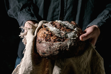 Man in black shirt holding in hands fresh baked artisan round homemade chocolate and cranberries rye bread on linen cloth.