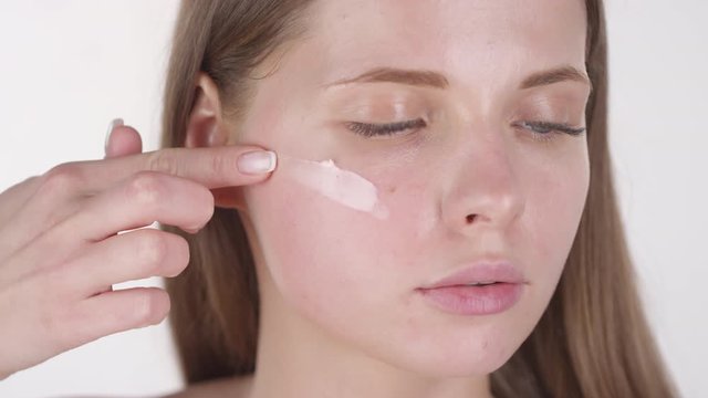 Close up shot of beautiful young woman putting dab of foundation on her cheekbone and smearing it, then looking at camera