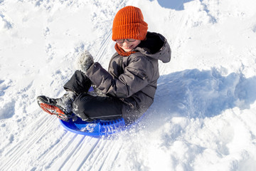 Fototapeta na wymiar A teenager flies on a round sled from a hill. White snow scatters to the sides. Winter games for children. A Smiling boy with spectacles on eyes in orange hat riding down with extreme speed.Sunny day