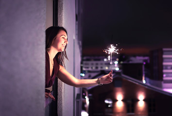 Sparkle in the night. Woman holding a sparkler out of window. New Year's Eve, Christmas party or birthday celebration at home. Happy elegant lady celebrating. City view. Light from firework stick. - Powered by Adobe