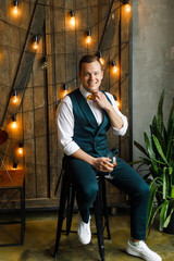 young man in a classic suit sits in a loft apartment on the background of wooden doors and light bulbs and drinks whiskey