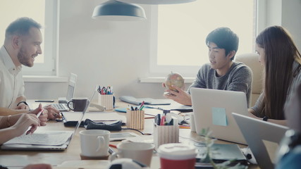 Happy Asian businessman takes a little globe, points at country at multiethnic office meeting in light loft coworking 4K