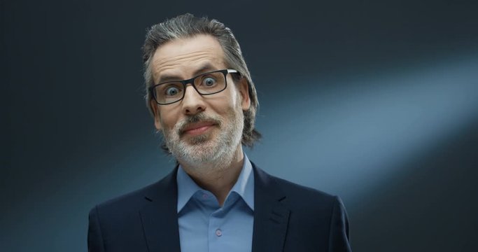 Caucasian businessman in glasses with gray beard tapping and texting on smartphone. Man in suit looking with big surprised eyes at camera on grey background. Senior male mobile phone user wondering.