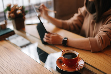 Close-up of a cup of dark coffee on a table in a cafe. In the background sits a unrecognizable young woman in casual clothes and works at a digital tablet.