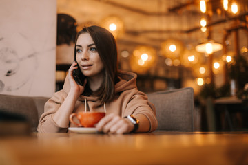 Middle shot portrait of young beautiful woman in casual clothes talking on the phone at a table in a cozy cafe. Coffee cup on the table. Concept of leisure activity.