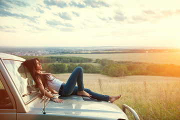Attractive yong woman is lying on the car's hood and looking at sunset. Rural evening background.