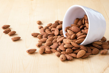 Closeup almonds nut are poured from white bowl on wooden table.