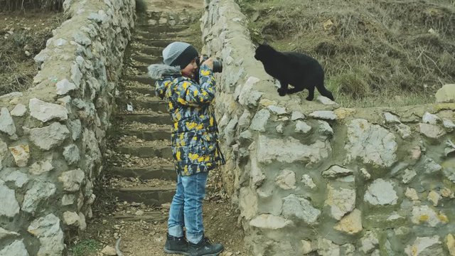 A boy in a jacket and hat takes pictures of a black cat sitting on a stone parapet