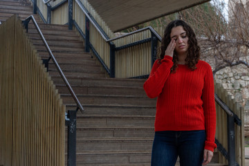 Portrait of tired attractive caucasian young woman model touching eyes, in the park, orange sweater and jeans, long curly hair. Place for your text in copy space.