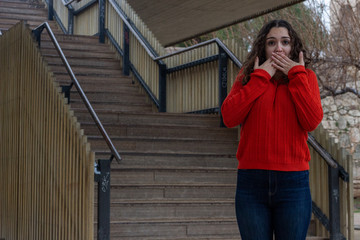 shocked scared attractive caucasian young woman model covering mouth with both hands, horrified stunned looking at camera isolated, in the park, orange sweater and jeans, long curly hair. Place for yo