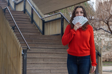 Portrait of attractive young woman with dollars in hands, banknotes, in the park, orange sweater and jeans. Place for your text in copy space.