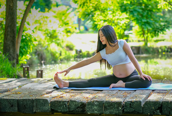 Portrait of young Asian pregnant woman exercise with yoga action by sit on wood bridge near the river in the garden with morning light.