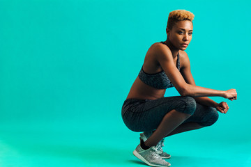 Portrait of a serious female athlete in sports bra and gym tights, squatting and looking off, against studio background