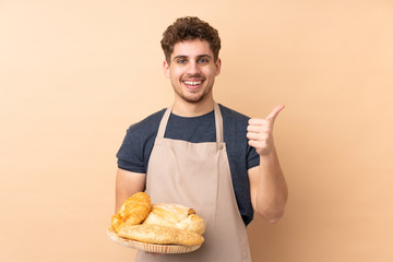 Male baker holding a table with several breads isolated on beige background giving a thumbs up gesture