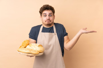 Male baker holding a table with several breads isolated on beige background having doubts with confuse face expression