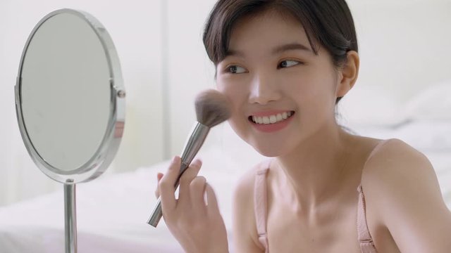 Beauty young asian woman happy and smile with face looking mirror applying makeup with brusher of cheek in the room, beautiful of girl holding brush make-up on facial, skin care and cosmetic concept.