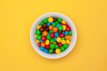 White bowl with chocolate colored round candy on yellow background in the center. Top view.