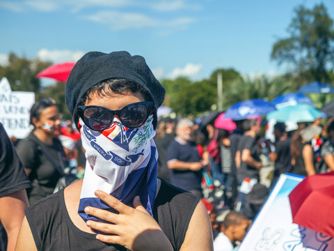 A girl with glasses, hat and scarf covering her mouth, fights for her rights with the Dominican flags and the blue sky in the background