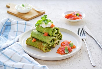 Spinach pancakes rolled up on a white plate on a light concrete background. Served with salted salmon and soft cheese. Spinach recipes.