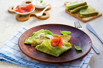 Spinach pancakes rolled up in triangles on a brown clay plate on a light concrete background. Served with salted salmon and soft cheese. Spinach recipes.