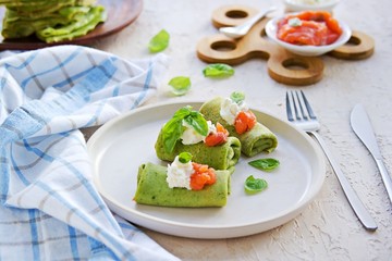 Spinach pancakes rolled up on a white plate on a light concrete background. Served with salted salmon and soft cheese. Spinach recipes.