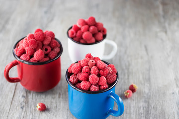 Organic raspberries in blue, red and white metal cups on gray wooden background.