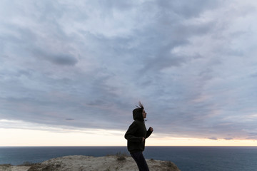 Girl runner is training on a high cliff above the sea. Morning jogging in nature by the ocean. Cardio exercise in sweatshirt tracking shot