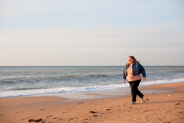 Beautiful overweight woman running on the sandy beach. Plus size girl enjoy warmth sunset with romantic mood. Fat model dressed jeans jacket and pink knitted sweater
