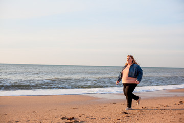 Beautiful overweight woman running on the sandy beach. Plus size girl enjoy warmth sunset with romantic mood. Fat model dressed jeans jacket and pink knitted sweater