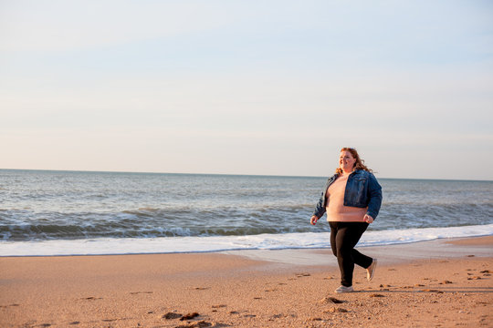 Beautiful Overweight Woman Running On The Sandy Beach. Plus Size Girl Enjoy Warmth Sunset With Romantic Mood. Fat Model Dressed Jeans Jacket And Pink Knitted Sweater