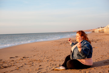 Fototapeta na wymiar Back view of beautiful overweight woman sitting on the sandy beach. Plus size girl enjoy warmth sunset with romantic mood. Fat model dressed jeans jacket and pink knitted sweater