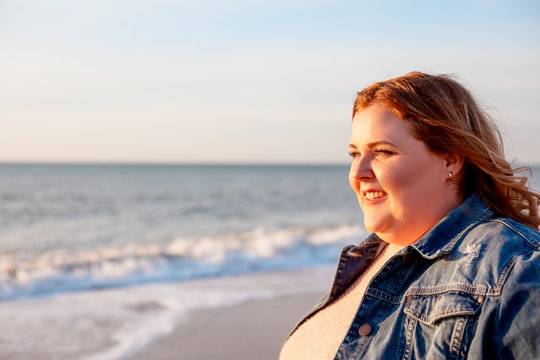 Back View Of Beautiful Overweight Woman Walking On The Sandy Beach. Plus Size Girl Enjoy Warmth Sunset With Romantic Mood. Fat Model Dressed Jeans Jacket And Pink Knitted Sweater