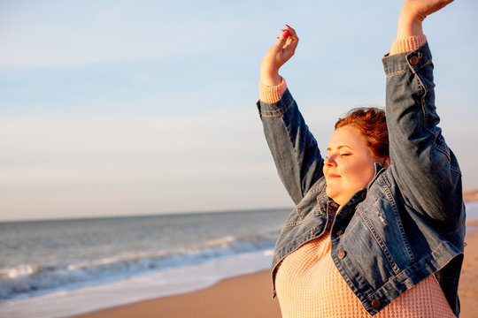 Back view of beautiful overweight woman sitting on the sandy beach. Plus size girl enjoy warmth sunset with romantic mood. Fat model dressed jeans jacket and pink knitted sweater