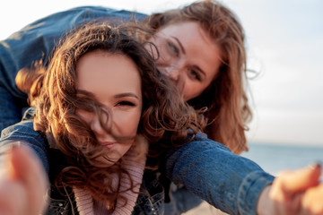 Back view of two freinds , plus size with  thin  girls walking on the fall  beach. Fat woman with strong friend  laughting and holding camera to do selfie. Overweight woman dressed jeans jacket
