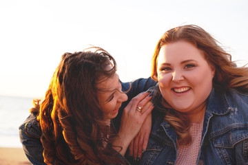 Back view of two freinds , plus size with  thin  girls walking on the fall  beach. Fat woman with strong friend  laughting and holding camera to do selfie. Overweight woman dressed jeans jacket 