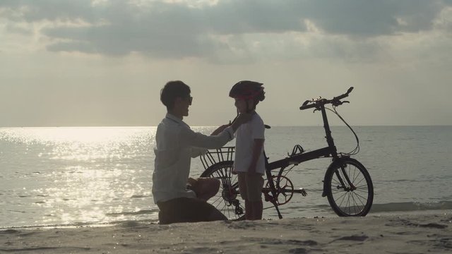 Happy asian family with father wearing helmet son and riding bicycle on beach in vacation together, parent and boy go to travel trip with cycling for fun and leisure in holiday, summer concept.