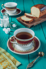 A tiffany color wooden table with a sliced cake on the cutting board, sugar cubes and two porcelain cups of tea. 