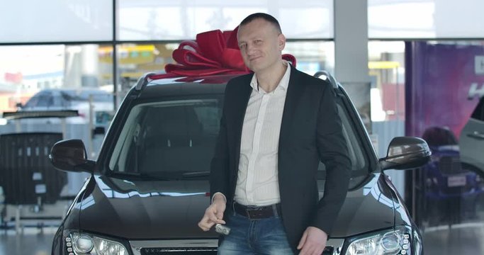 Portrait of confident Caucasian businessman bragging car keys and crossing hands. Young successful man standing in front of new vehicle in dealership and smiling. Cinema 4k ProRes HQ.