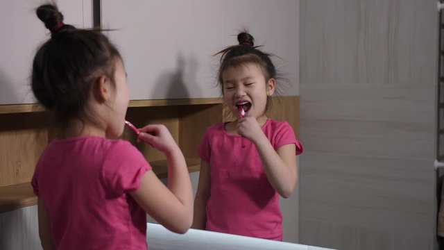 Back view of adorable chinese schoolgirl standing in front of mirror and thoroughly brushing her teeth in bathroom. Morning family rituals, dental care and caries prevention from early age concept