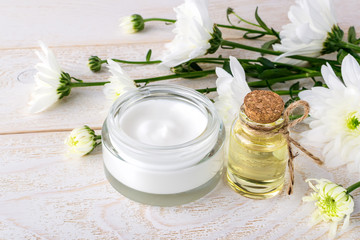 Obraz na płótnie Canvas Facial cream in an open glass jar, chrysanthemum essential oil in a glass bottle and white chrysanthemums on a wooden surface. Spa, beauty, skincare and cosmetology