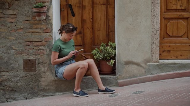 Young woman in sunglasses, denim shorts sits with smartphone at wooden doorstep