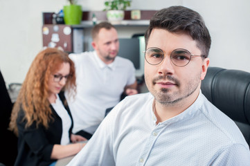 Portrait of young office worker in glasses sits with sitting colleagues in the background