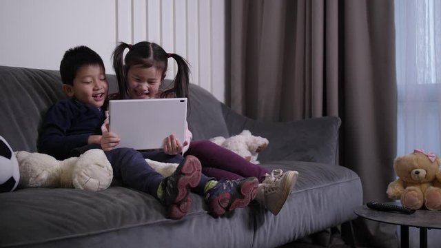 Cheerful little asian children laughing while watching funny video on tablet pc at home. Cute preadolescent siblings having fun during comedy movie while spending free time together in living room