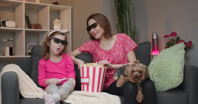 Woman with a little girl and dog Wearing 3d Glasses watching tv and eating popcorn. Family time relax with young girl kid on sofa in living room concept